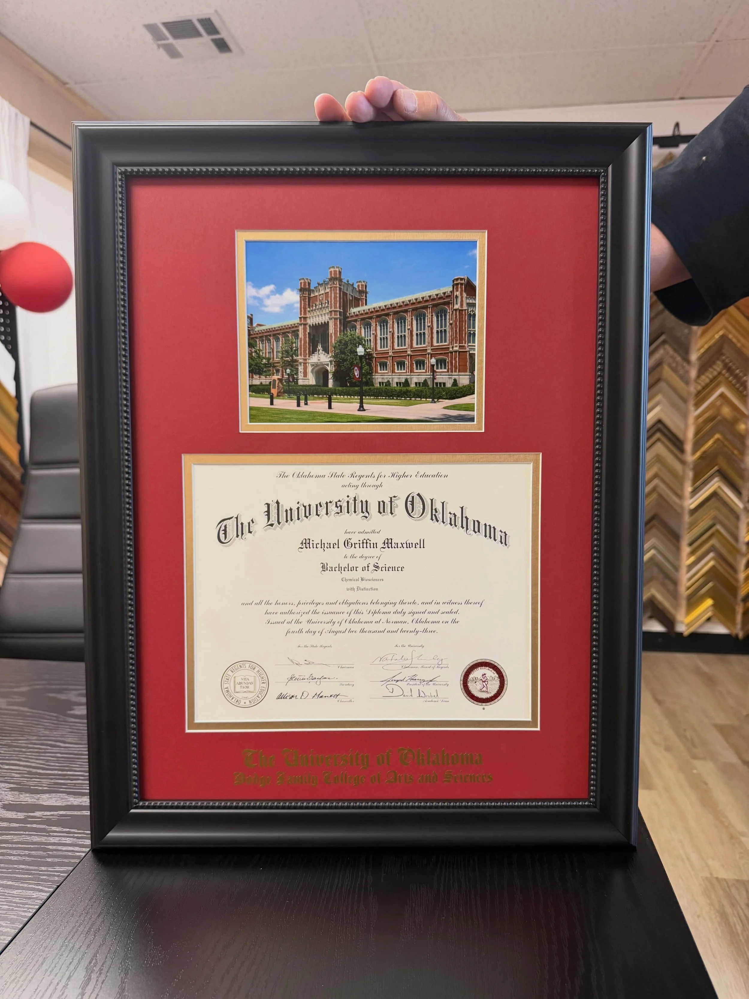 University diploma framed with a crimson and gold double mat alongside a campus photograph
